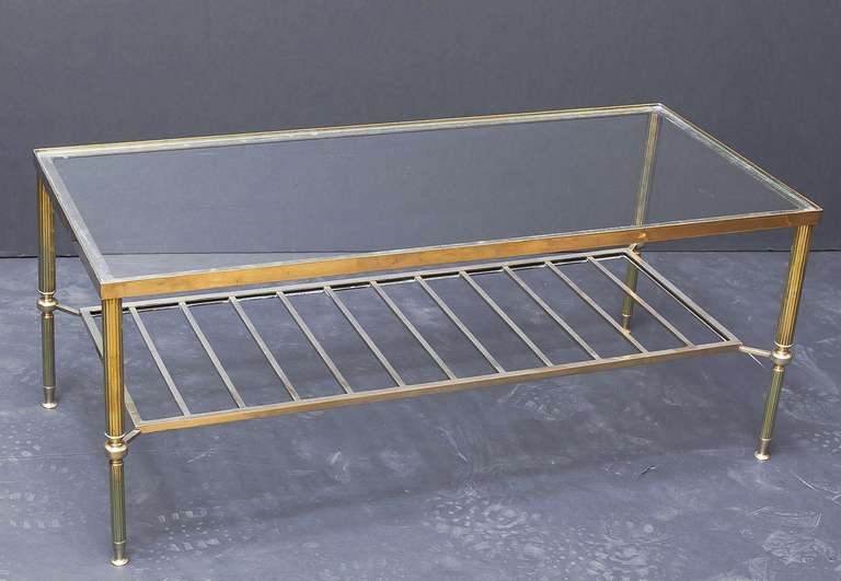 An English cocktail or coffee (low) table of heavy brass in a modern style featuring a rectangular clear glass top over reeded brass legs with a magazine rack lower tier.
