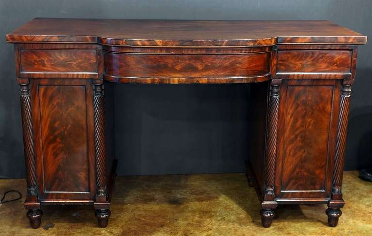 A handsome English breakfront pedestal sideboard or serving console of flame-cut mahogany, featuring a moulded top over a frieze with two short drawers flanking a fitted breakfront center drawer, set upon two pedestals, each with door with two