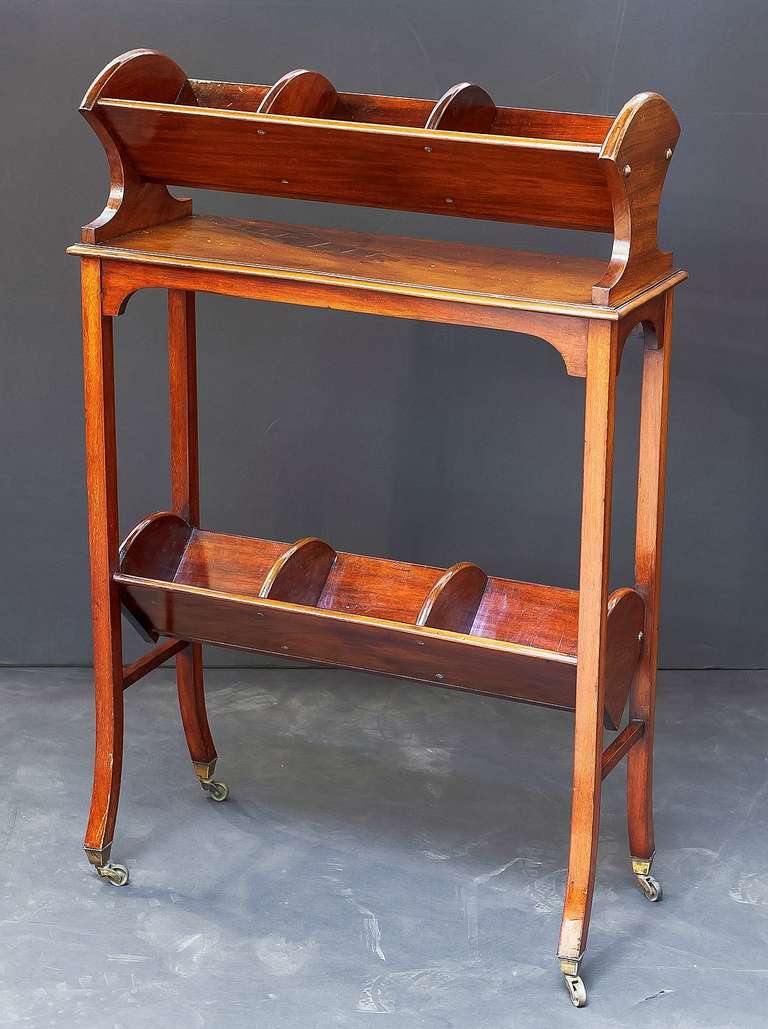 A handsome Scottish two-tiered book stand or library stand of mahogany, featuring a canted upper tier with three sections for octavo-sized books, attached to a bottom tier with similar three section canted shelf, and resting on rolling brass casters.