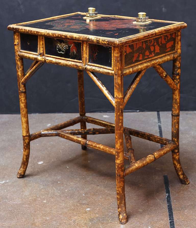 An English bamboo desk with lacquered top and fitted inkwells, over a frieze with lacquered panels and one drawer, on bamboo legs.