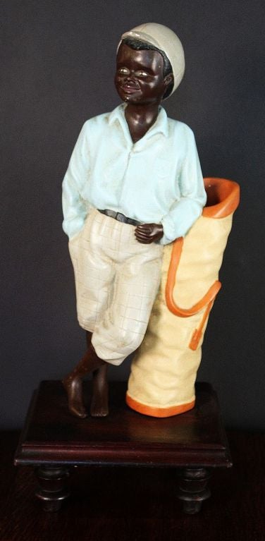 A vintage painted chalkware figure of a black golf caddy (or caddie), sporting a cap and leaning on a golf bag, mounted to a moulded square base of mahogany with turned supports.<br />
A timeless example of Golfing memorabilia.