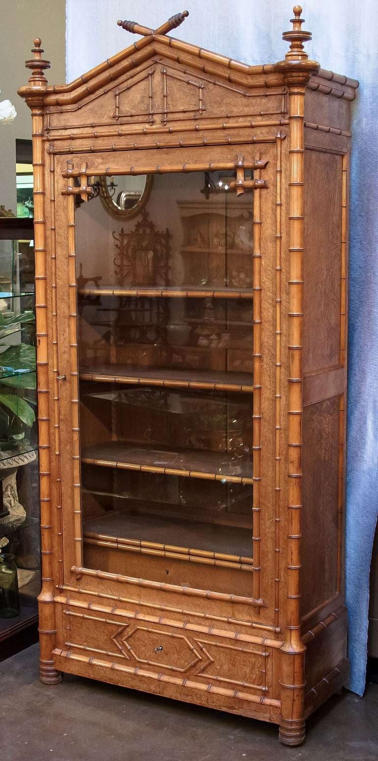 A handsome faux bamboo armoire (or bookcase and display cabinet) of curly maple featuring a canopy top with decorative finials, over a glass or glazed door enclosing a cupboard space with adjustable shelves, each with faux bamboo accents. 
The base