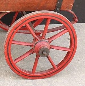 An English goat cart featuring a carry-all of painted wood with railing attached to an iron under-carriage set upon four spoke wheels of painted wood, designed to be pulled by the wooden harness attachments at the front of the cart.