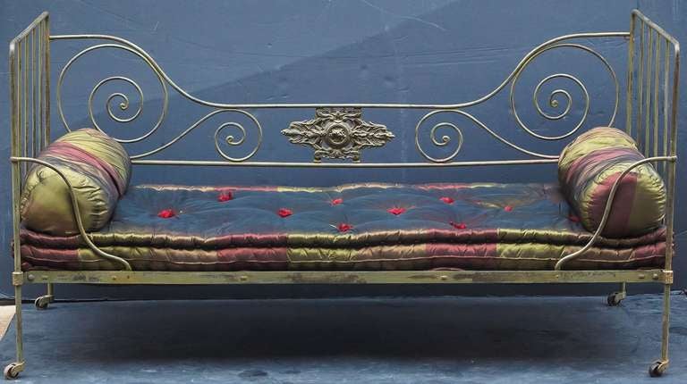 A period French day bed of painted wrought iron featuring a scroll design to the back with a central decorative cartouche, with latticework seating supporting upholstered bedding and two roll cushions. Resting on casters.

H 37