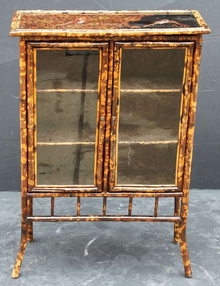 An English bamboo bookcase featuring a japan-lacquered top with bamboo accents over two framed-glass doors with brass hardware, opening to an interior of two bamboo-edged shelves, each lined with pressed paper, and resting on bamboo legs.