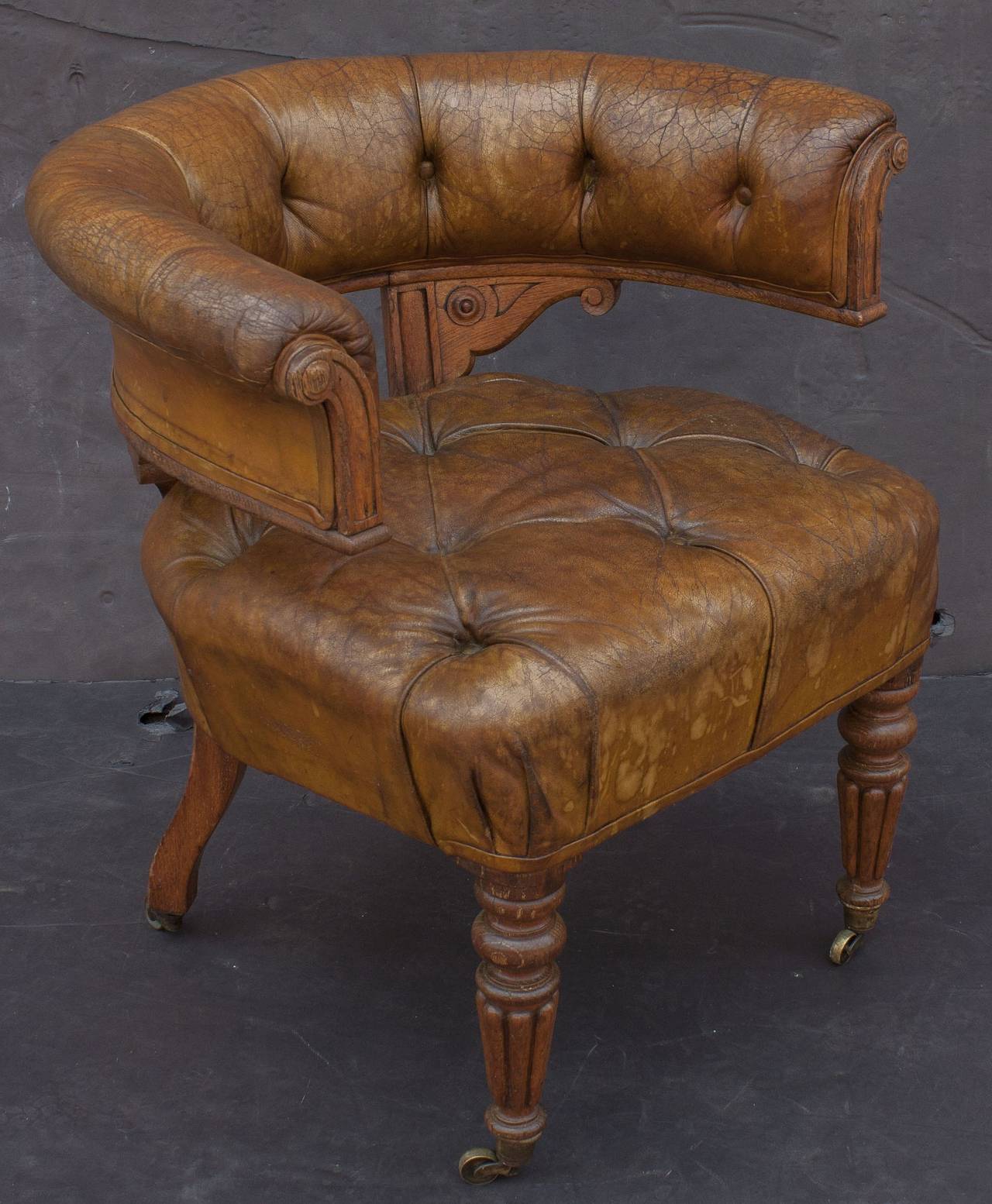 A fine English desk chair of tufted leather and oak on rolling casters, featuring a roll-back arm with tufted-leather padding, mounted to a tufted-leather cushion seat and set upon a frame of turned legs.