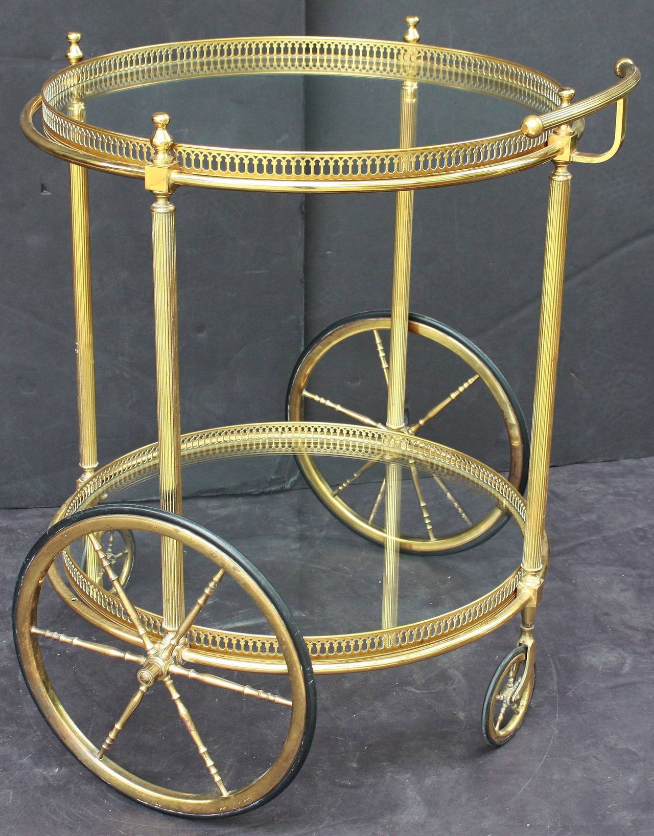 A handsome French rolling drinks cart or trolley (bar cart) of brass and glass, featuring two round tiers, each with pierced gallery and glass, mounted to a frame with curved handle, finials, and four brass spoked wheels - Two large fixed wheels and