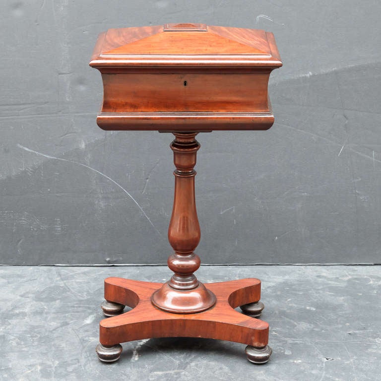 An English teapoy (or tea caddy on stand) of mahogany, featuring a hinged-lid box set upon a a column post stand with platform base, the interior with three compartments for storing tea. With key.