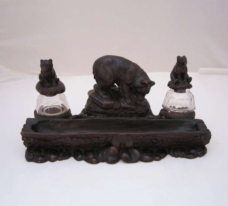 A blackforest desktop ink well set featuring a large carved bear in the center, opening as a hinged lid for pen nibs, flanked on opposing sides by faceted, fitted glass inkwells, each capped by a carved bear cub top, with pen holder tray  to front