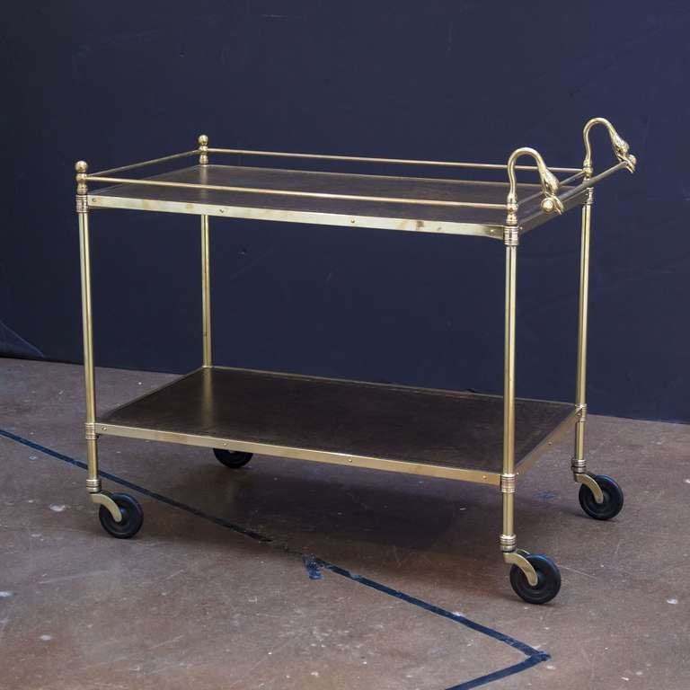 A handsome two-tiered drinks cart or serving trolley (bar cart) featuring a swan's neck design on the handle. The top tier with brass gallery railing and finials, each tier with embossed leather top, resting on rolling caster wheels.

Makes a