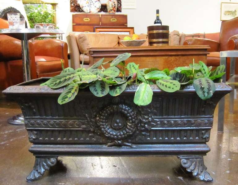 A French low planter or rectangular urn of cast iron featuring a decorative neoclassical design with center relief of a wreath on opposing sides, resting on acanthus leaf feet.