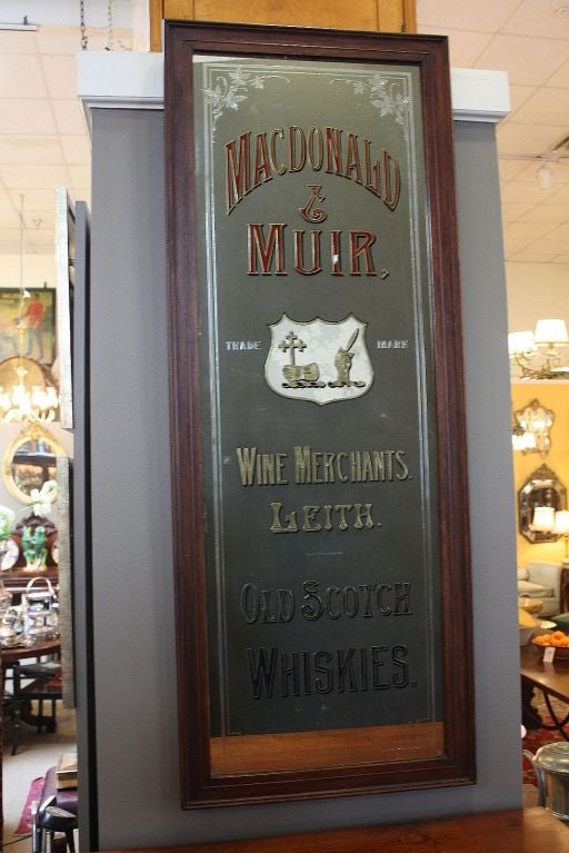 A vintage Scotch whisky advertisement (portrait) mirror (or artistic show card), framed in mahogany and featuring an etched and painted mirrored glass reading: <br />
Macdonald and Muir. <br />
Trade Mark. <br />
Wine Merchants. <br />
Leith.