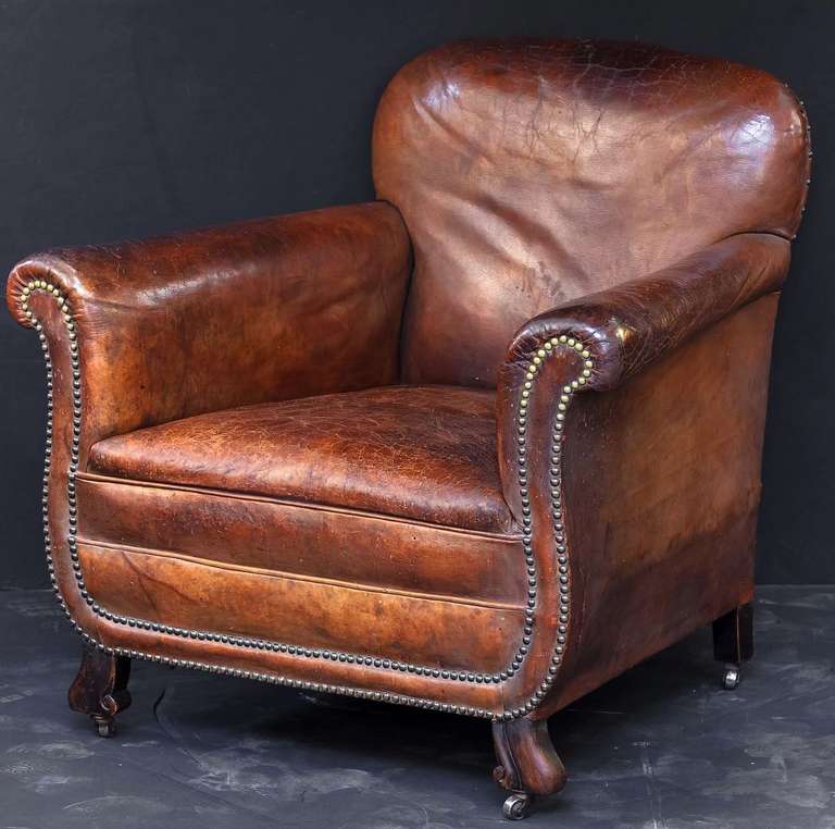 A handsome French upholstered leather club chair featuring scroll back and arms with nail-head trim and set upon stylised, turned wood feet with casters.