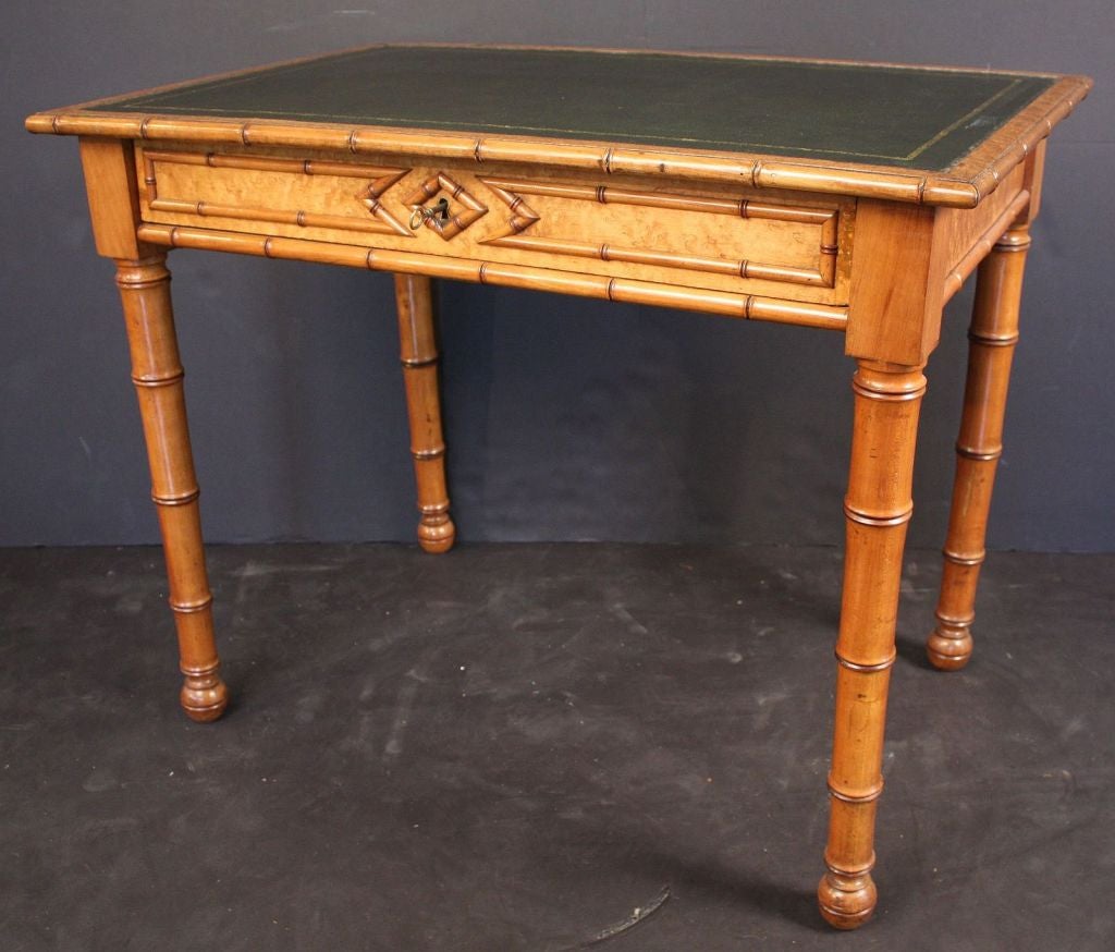 A Faux Bamboo writing table or desk in maple featuring a moulded top with embossed leather writing surface and faux bamboo design around the circumference, over a frieze of curly maple with locking long drawer (with key), and resting on four carved