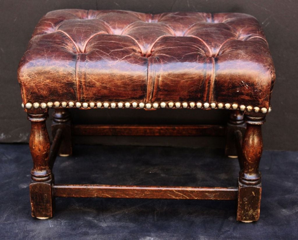 An English footstool in the Chesterfield style, featuring an upholstered top of button-tufted leather and nailhead trim around the circumference attached to a nicely-turned wooden support.