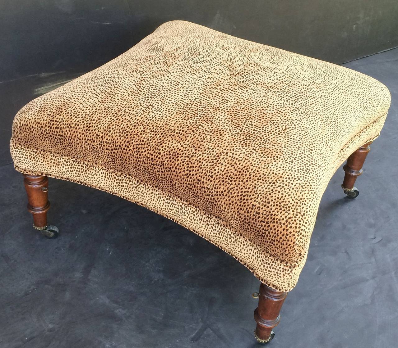 A handsome English upholstered ottoman (or stool) featuring a leopard print fabric on a wood frame with four turned mahogany legs, each with rolling brass casters.