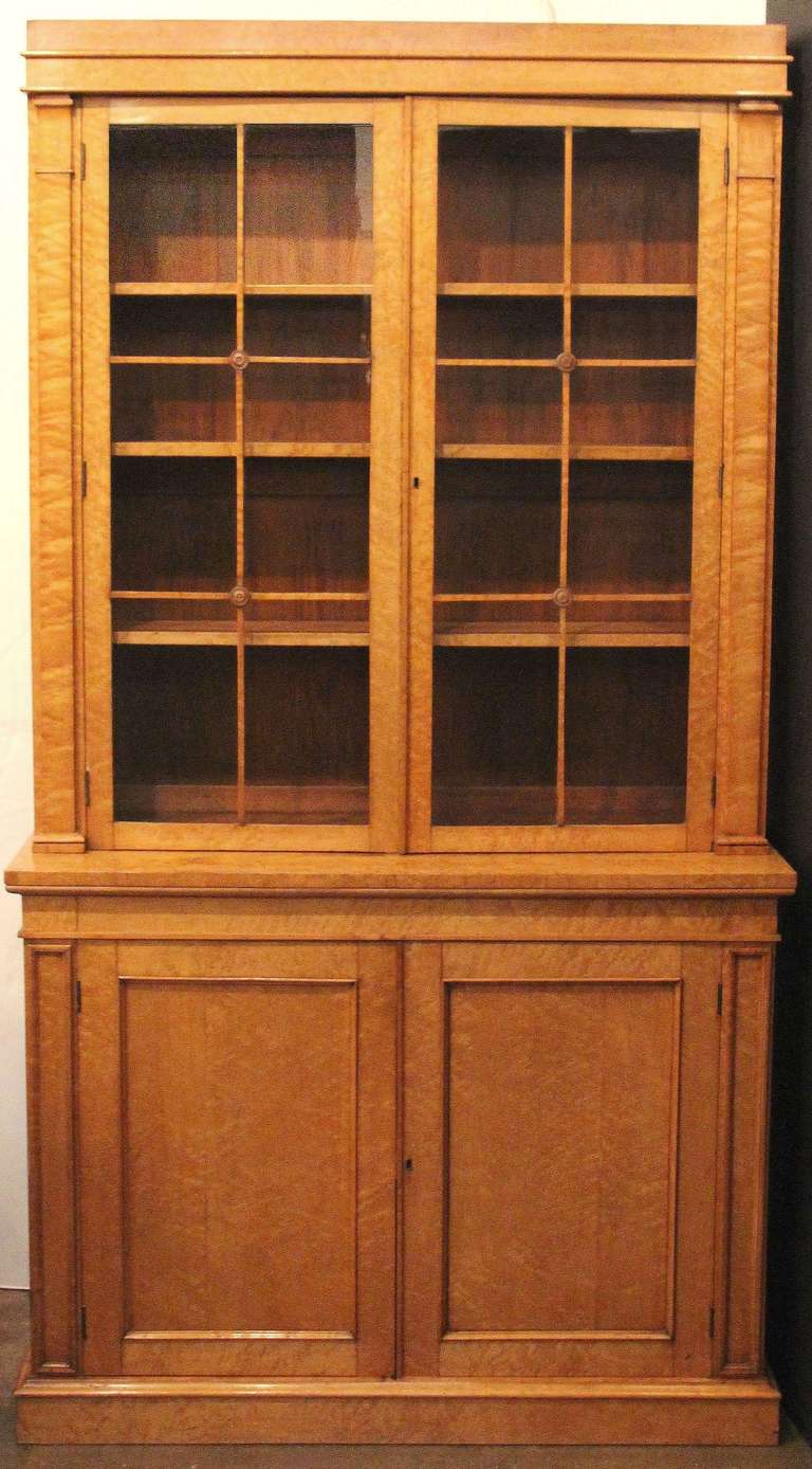 A handsome large two-tier bookcase featuring a curly maple exterior in the Biedermeier style. The top tier showing two glazed doors with escutcheon and key flanked by two raised columns of curly maple, the interior with three drawers, and crowned by