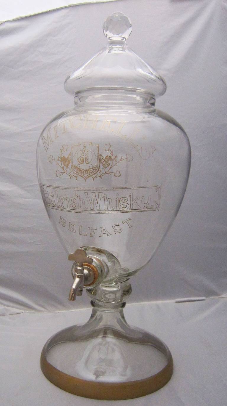 A large bar-sized footed glass whiskey dispenser (or decanter) with removable top and working spigot. With gilt painted accents on the body.

Marked: Mitchell's Old Irish Whisky - Belfast