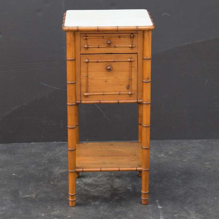A French faux bamboo nightstand or bedside table (petite cabinet or commode) of long-leaf pine, featuring a white marble top over a frieze with drawer and cabinet fall attached to four turned legs and bottom tier. The whole accentuated by a faux
