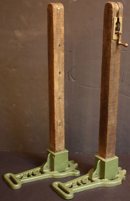 An authentic pair of English tennis posts featuring heavy cast iron bases (painted green), each set with rounded end posts of wood. One post with eye hooks for netting, the other with eye hooks and winding lever (tennis net tensioner) with pulley