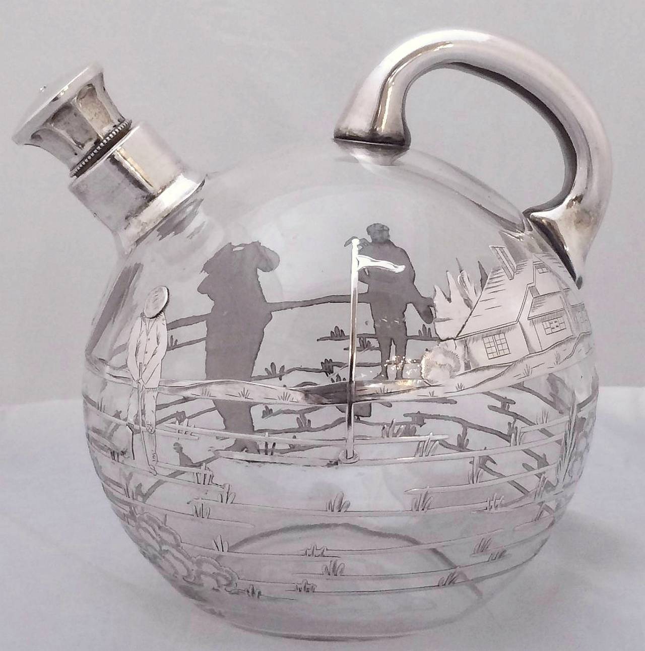 A handsome round English spirits decanter or claret jug featuring a glass body, handle, and spout with silver overlay. The silver overlay around the body featuring a design of two golfers on the obverse and a golfer with putting green,  flag, and
