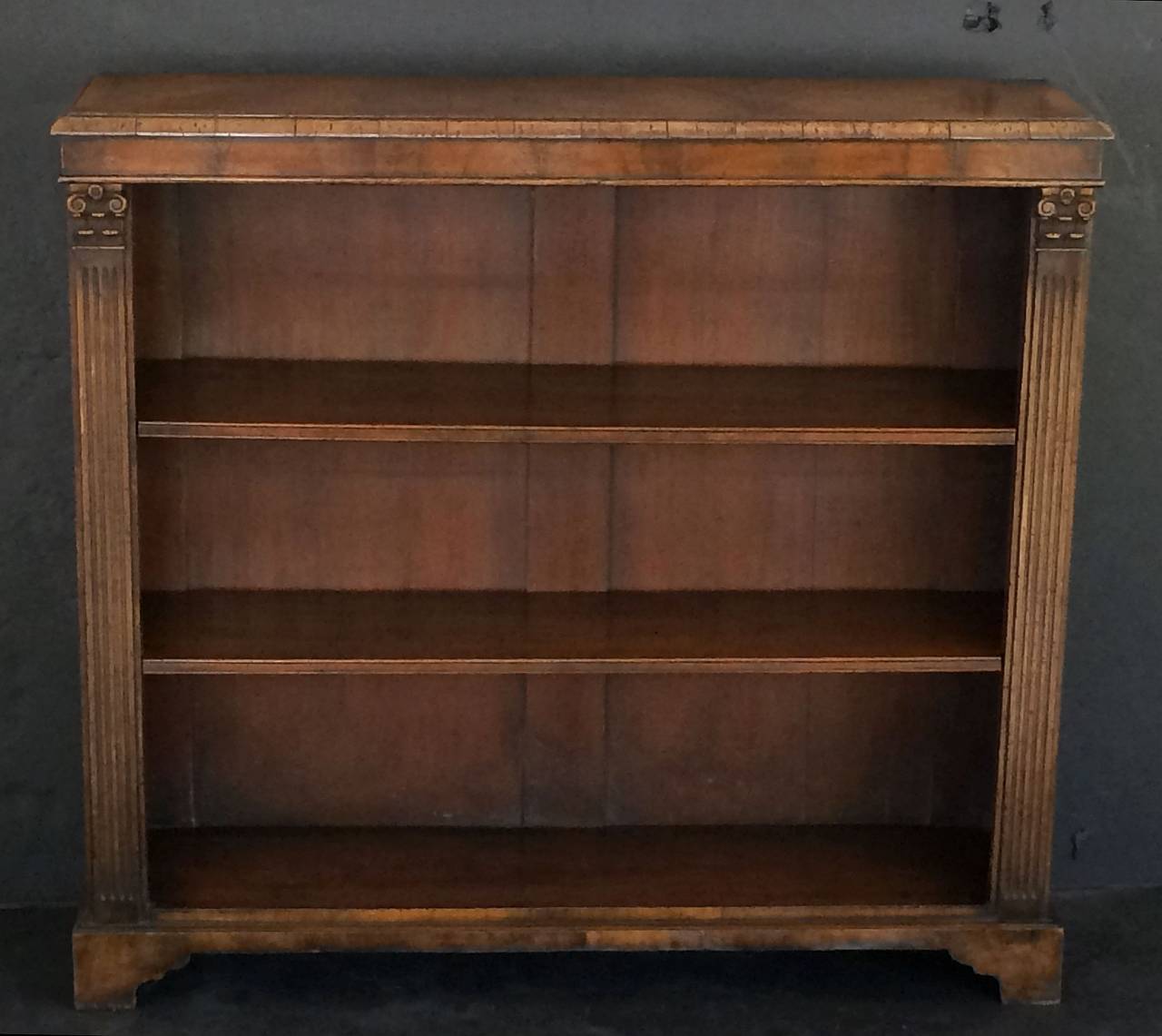 A fine English open bookcase of figured walnut, featuring a moulded top upon a case with two carved Classical reeded column sides, two adjustable, removable shelves, and set upon bracket feet.