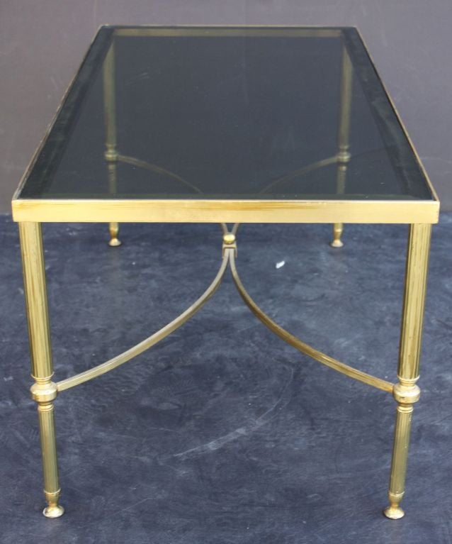 A mid-century period cocktail or low table of brass and rectangular smoked glass - the table on four reeded, tapering legs, adjoined to a bowed stretcher with finial.

Perfect for use as a coffee table.
