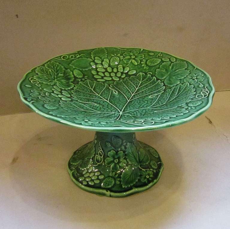 A fine English green-ware Majolica tazza or footed bowl featuring a green glaze pattern of a grape leaf with flowers and fruit on the top and column base.

Unmarked. Probably by the Wedgwood pottery.
Similar examples are pictured on pp. 126-127