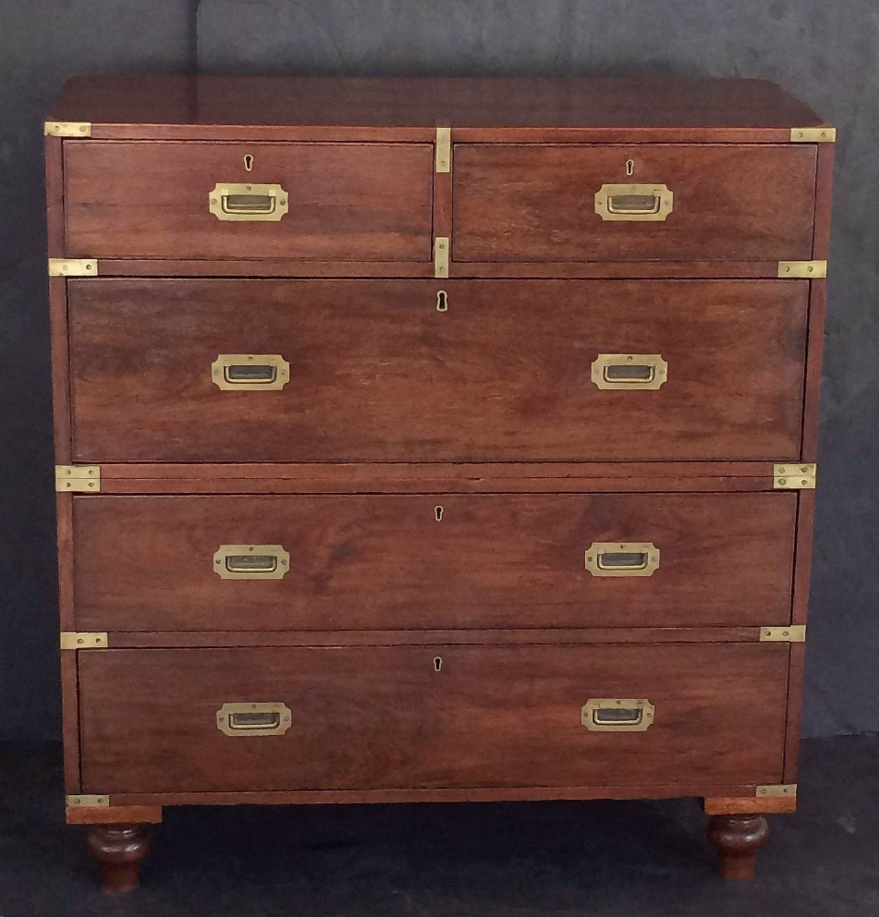 An English military officer’s Campaign ware chest, featuring a dark teakwood exterior, showing two short drawers over three long drawers. The brass-bound chest, in two parts, accented with recessed brass hardware, resting on shaped feet.

This