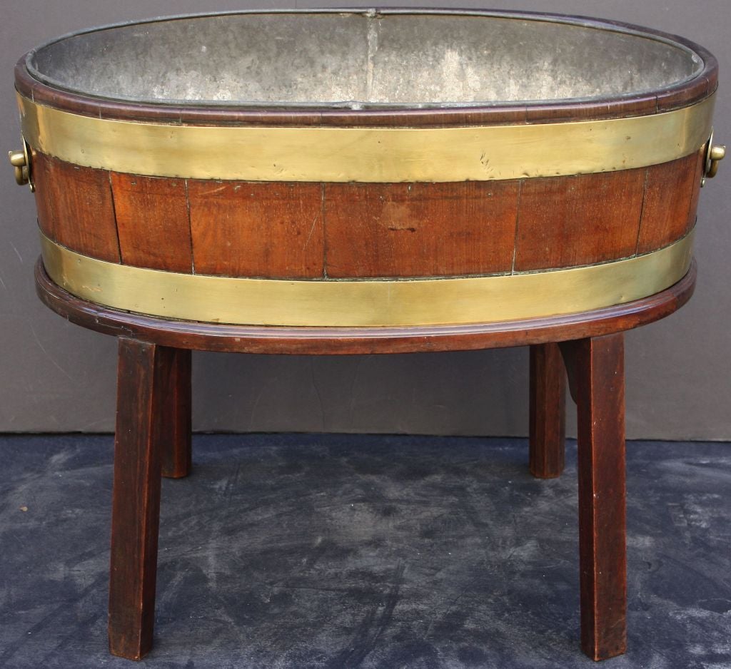 A handsome Georgian-era oval wine cooler on stand from Ireland, featuring a wine cooler of brass-bound wood with heavy brass handles, removable fitted zinc liner and fitted oval four-legged stand.