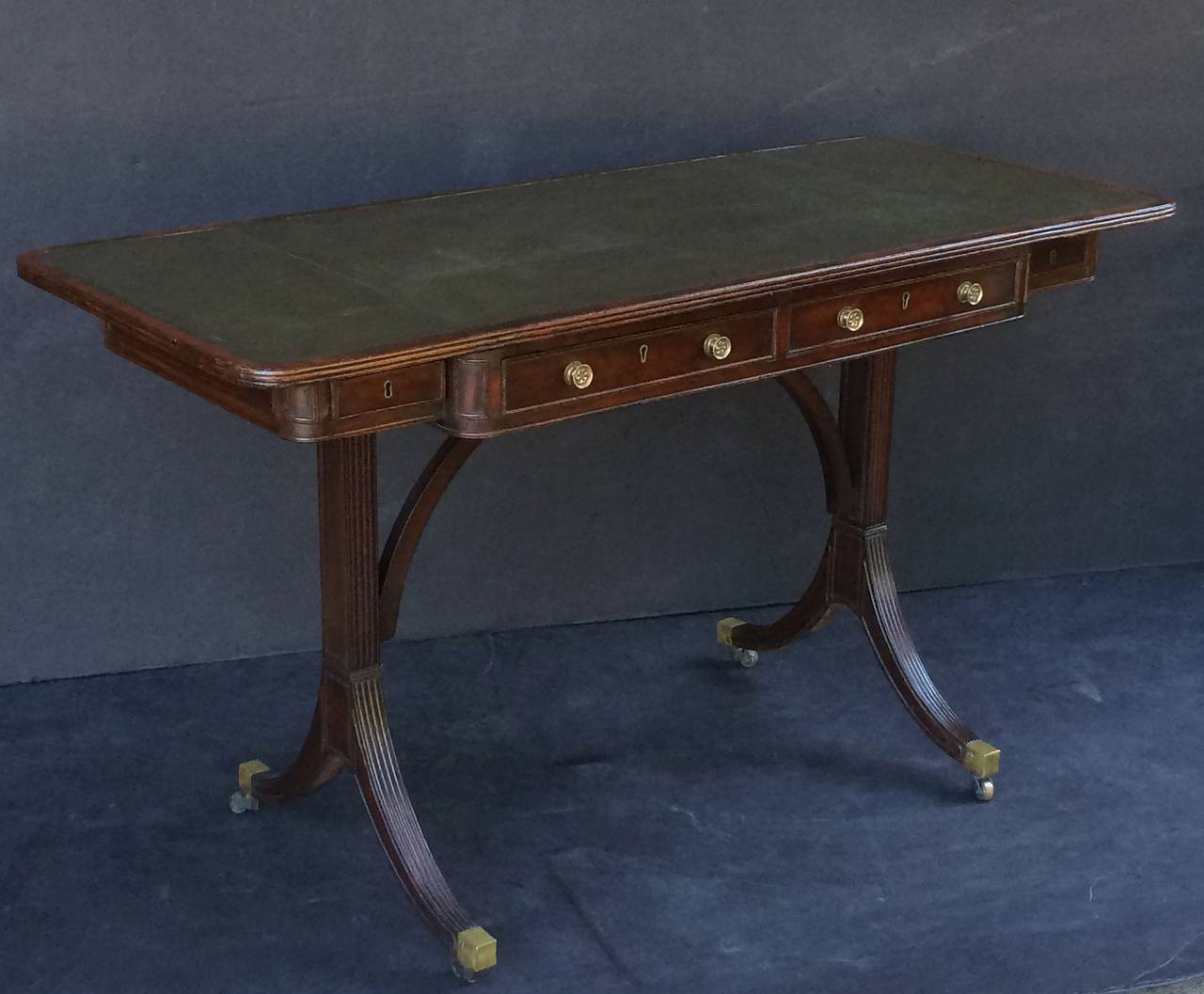 A fine English writing table or desk from the Regency period, featuring a moulded top with embossed leather, Over a breakfront frieze with four drawers, the opposing side with matching faux drawers. Resting on a reeded leg stretcher base, with brass