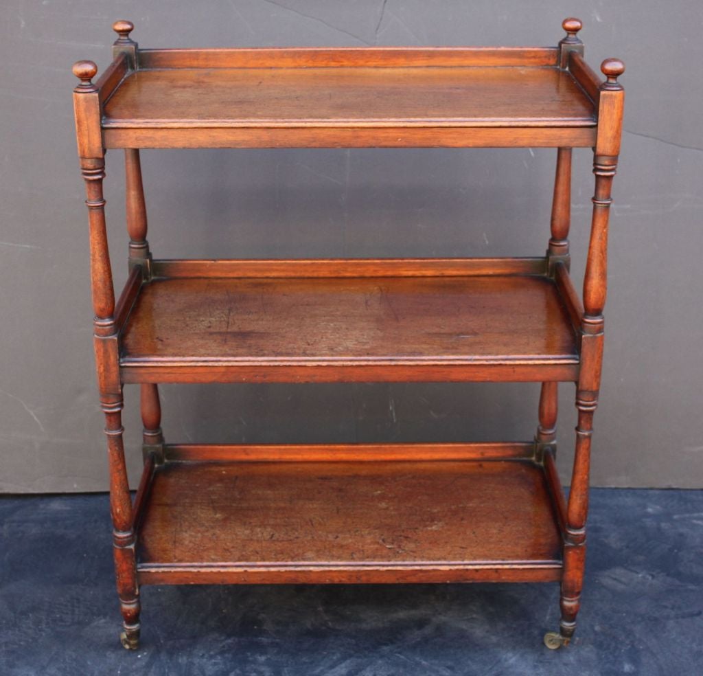 An English three-tiered trolley server or serving cart of mahogany featuring three moulded shelves (each with three quarter gallery) adjoined to four turned baluster supports, topped with finials and resting on rolling brass casters.