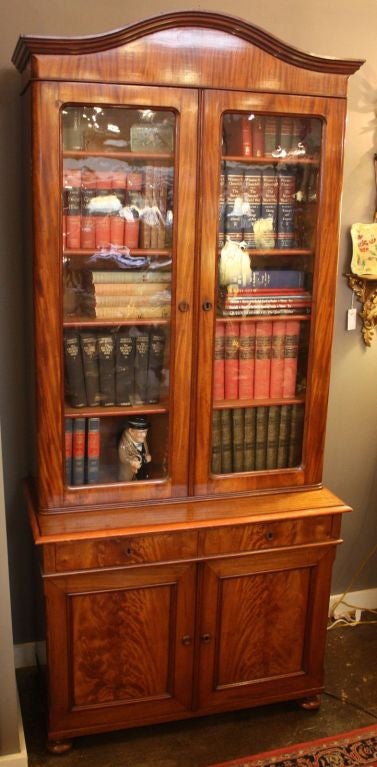 A fine Scottish two-tier bookcase cabinet of mahogany, featuring a canopy top over two glazed locking doors with key, opening to four adjustable shelves, over a bottom tier with two short drawers over two framed cabinet doors of flame-cut mahogany,