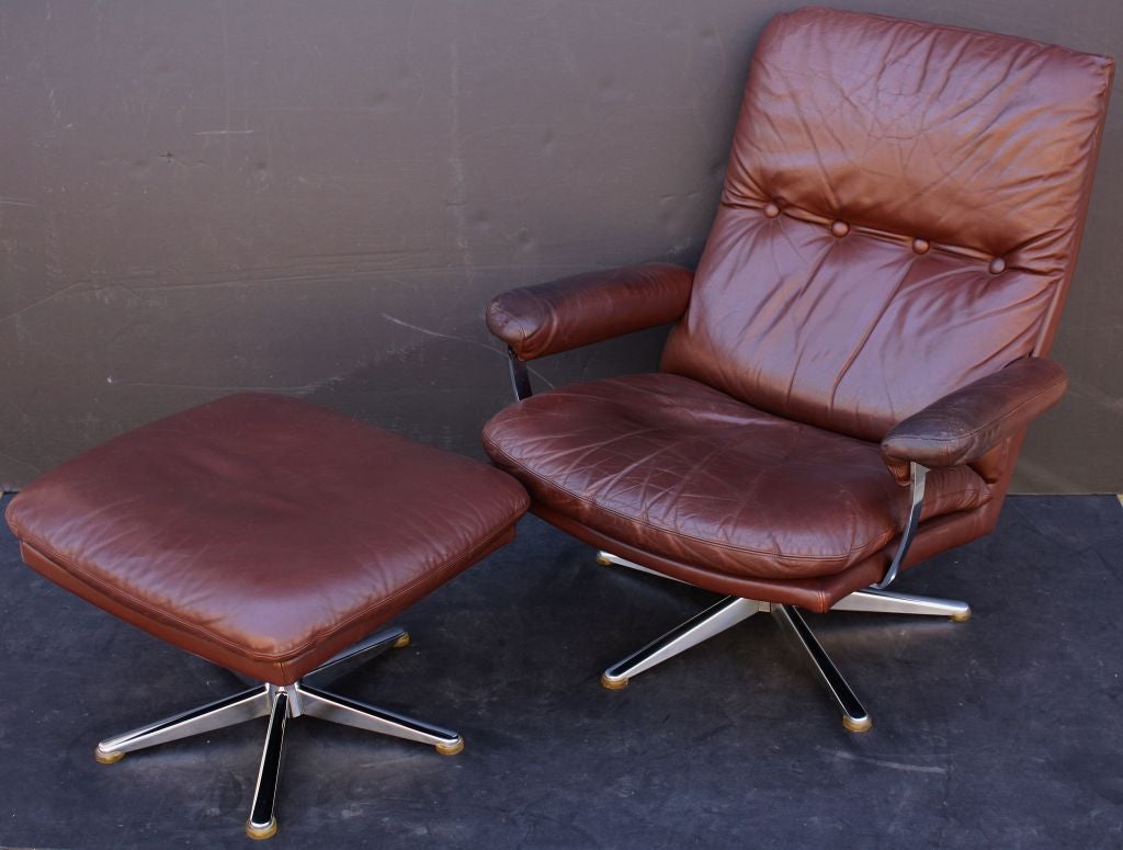 A Danish Modern swivel armchair recliner of leather with matching swivel stool or ottoman of leather. The Eames-style chair featuring a button-tufted back with padded seat and arms on a chromed steel five-legged pedestal base. The stool with similar