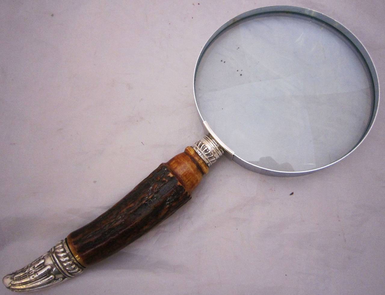 A large English hand magnifier or table top and desk magnifying glass featuring a large round looking glass mounted to a horn handle and capped with a silver foliate design. Round glass is 4 1/2