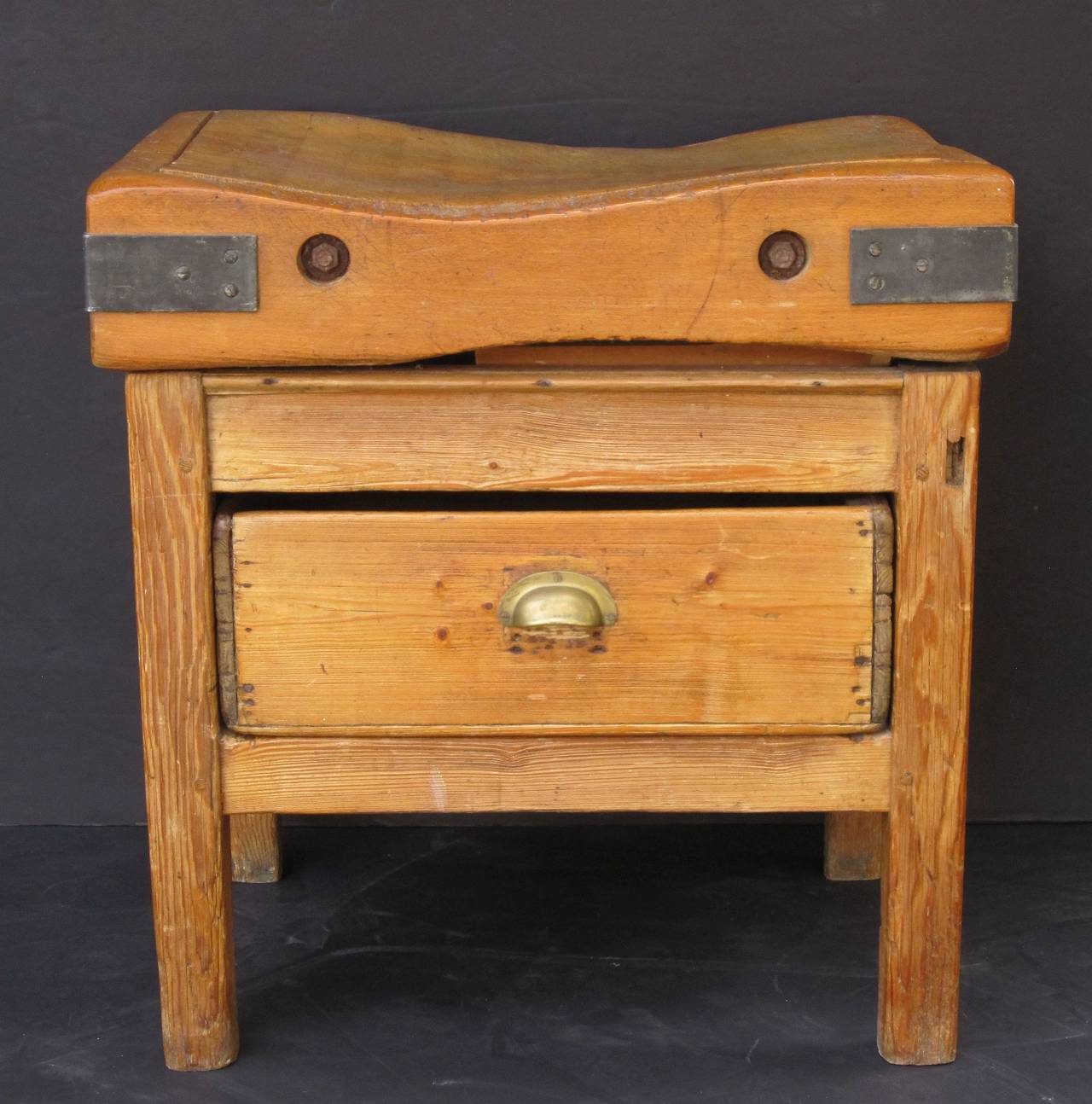 A two-tiered butcher's chopping block or table featuring a large, rectangular sloping block or slab of iron-bound wood set upon a bottom tier four-legged, paneled support stand of pine, with fitted drawer and brass pull handle, the drawer opening on