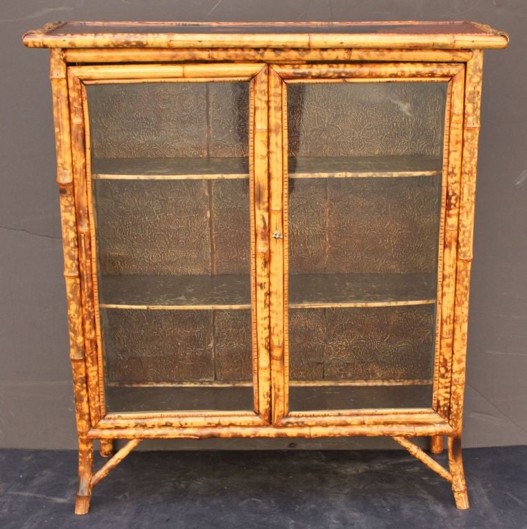 An English bamboo bookcase featuring a japan-lacquered top with bamboo accents over two framed-glass doors with brass hardware, opening to an interior of two bamboo-edged shelves, each lined with pressed paper, and resting on bamboo legs.
