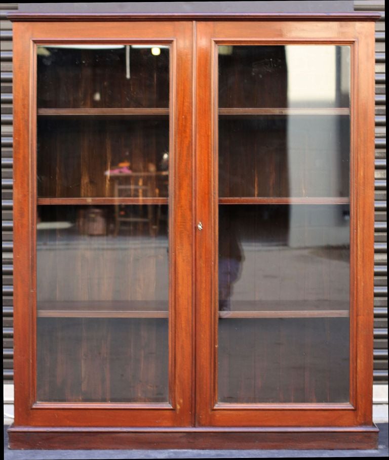 A fine English standing bookcase of mahogany featuring a moulded top over two framed glass doors, with lock and key, enclosing three shelves, set upon a plinth base.