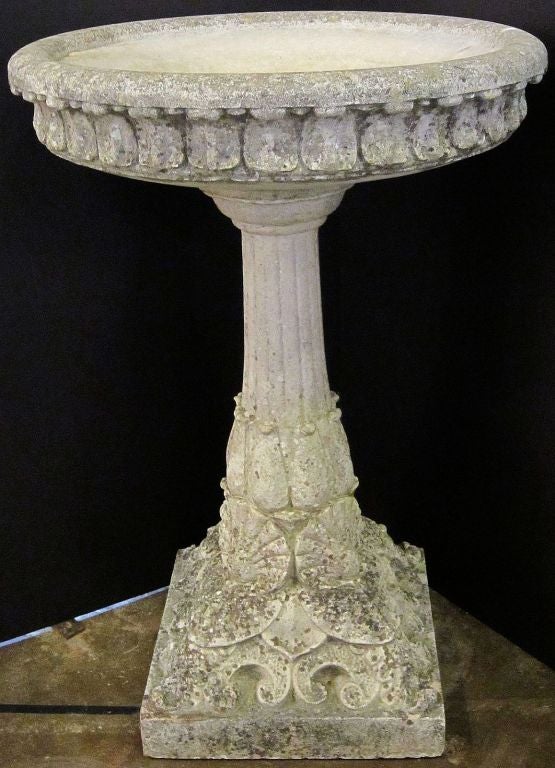 A large English bird bath of composition stone featuring a large diameter top with a Classical design of acanthus leaves around the circumference, over a column pedestal, with a shell and scroll design on the raised plinth base.

Perfect for a