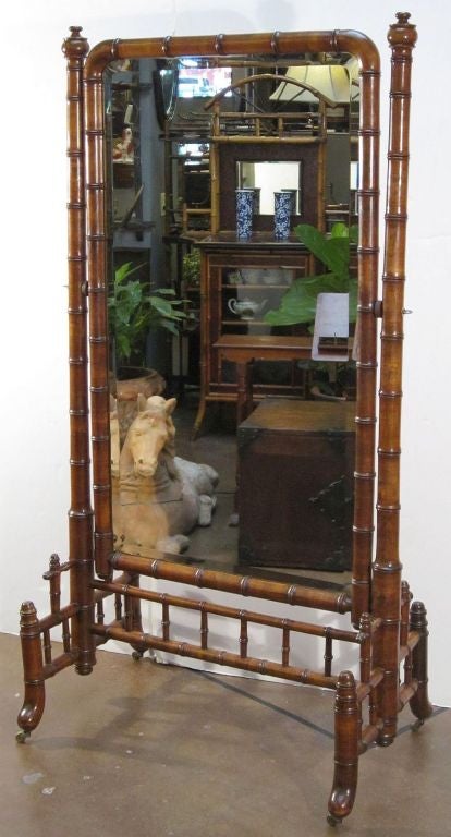 A faux bamboo cheval mirror featuring a mirrored glass panel framed in turned wood with a bamboo design mounted to a rolling frame of turned wood, capped with turned finials, with a stretcher of turned spindles above swept legs with casters.