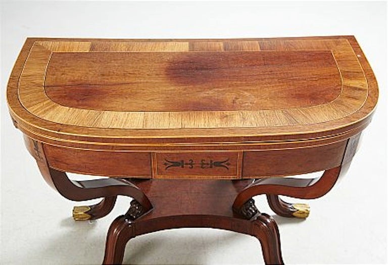An empire game table from France made during the Empire Period ca 1800. Veneered in mahogany and decorated with inlays and brass detailing. Opens up to a rectangular top. 