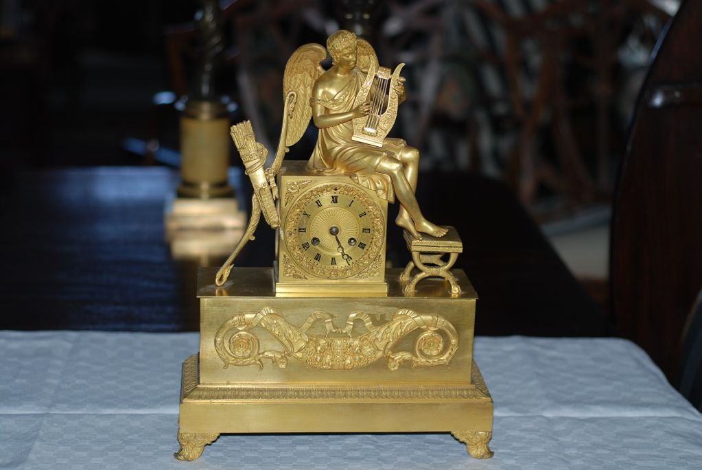 Clock Mantle Gilt Bronze Neoclassical Empire French 19th Century France. A French mantel clock in gilt bronze. Neoclassical symbols such Cupid playing a lyre. Next to him are his bow and arrows of love.