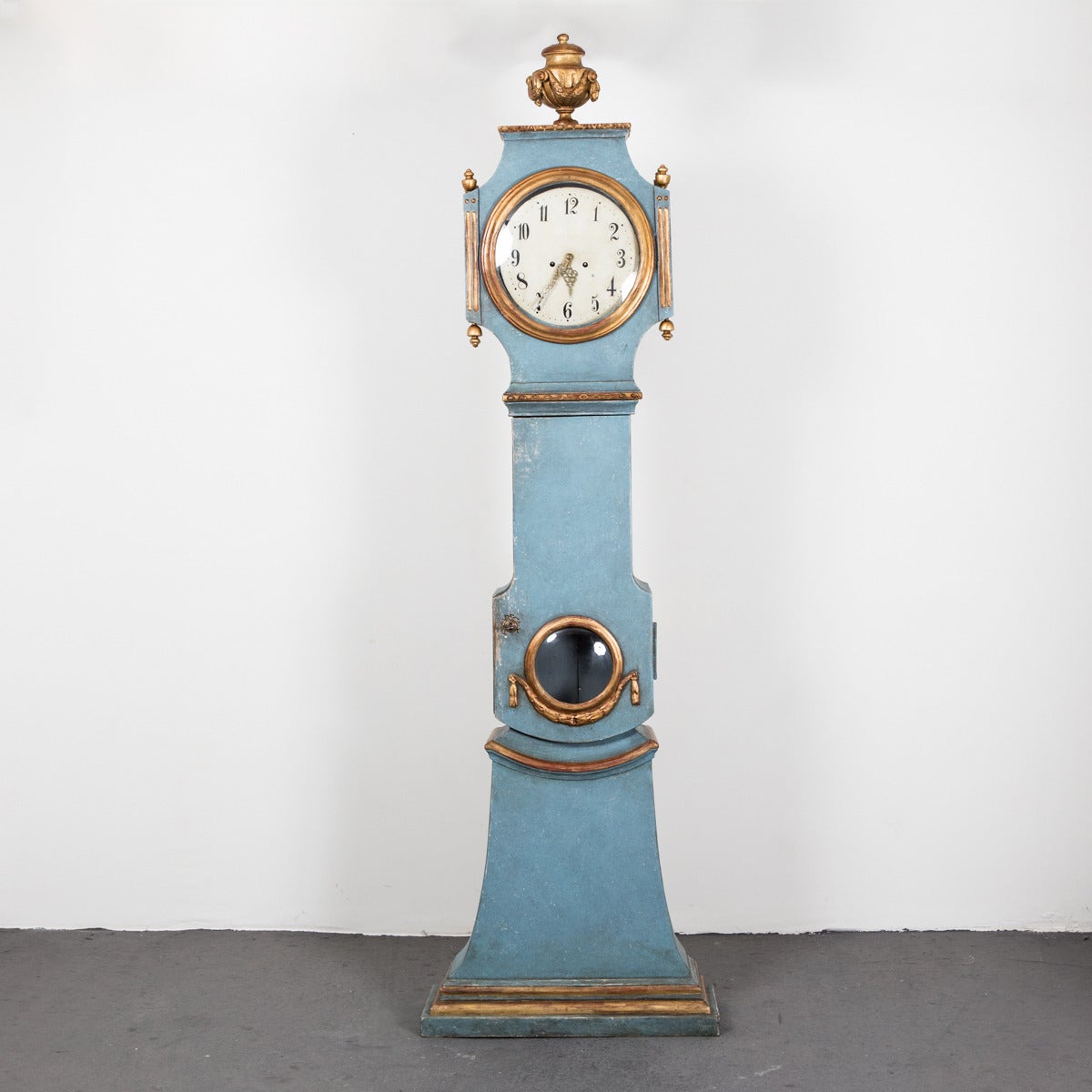 A blue painted Gustavian floor clock with gilt wood details. Top with a flowers swag decorated urn. Beautifully painted face and carved brass hands.