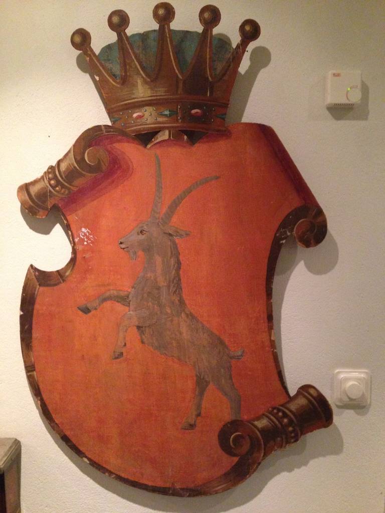 A shield made for a noble family's crest. Picturing a prancing aries with a crown on top. Painted with tempera colors on wood. Made in Sweden during the 19th century.