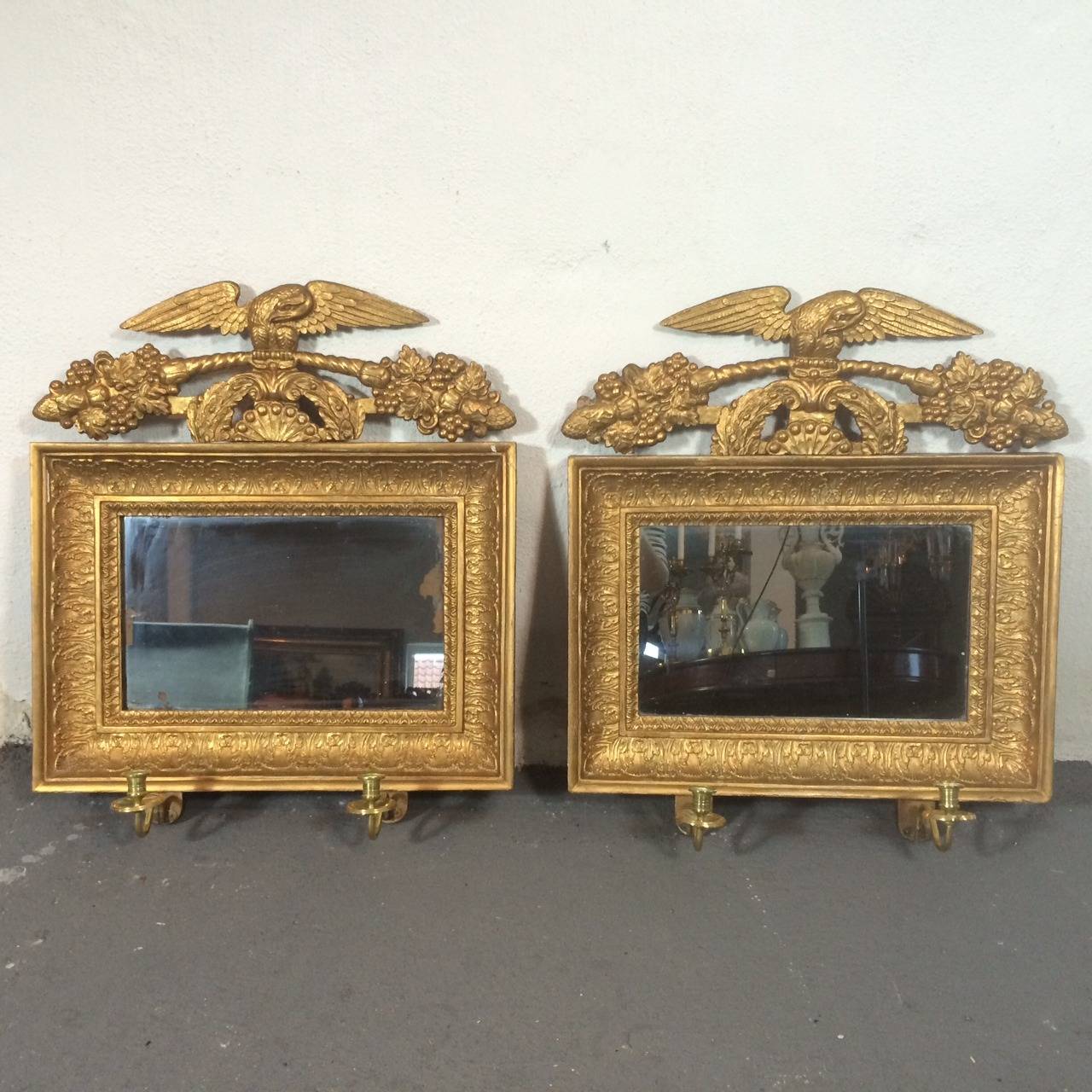 A pair of Swedish Neoclassical mirrored wall sconces in a gilded frame with carved decorations with an eagle, flowers and spears. Holders for 2 candles in brass on each sconce. Original mirror glass.