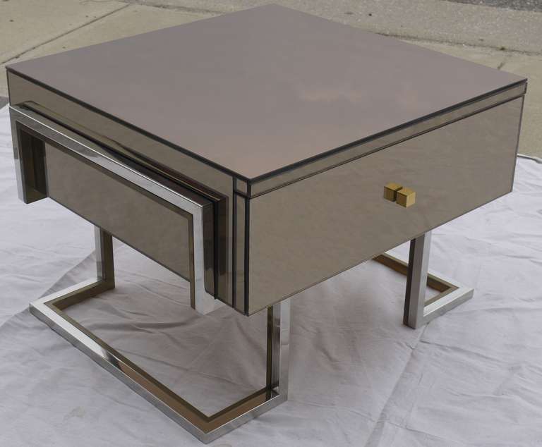 Stunning pair of mirrored side tables by Michel Pigneres in a signature smoky mirror covered table supported by a gilt brass and stainless base which wraps around and holds the drawer.  Beautiful yet simple design.  We have two pairs available,