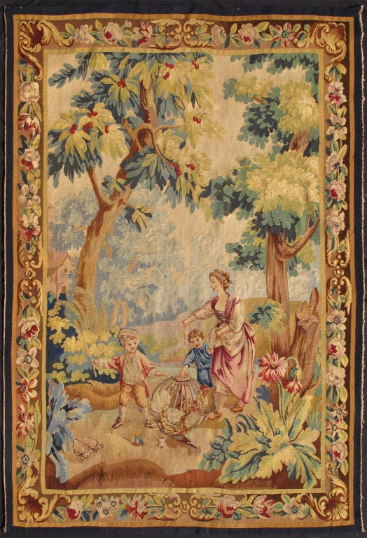 Delightful in both subject and palette, this Antique Verde Tapestry brings to life a scene from the 19 century, showcasing a mother with her two children playing within an enchanting woodland setting. The entire scene is framed by a highly