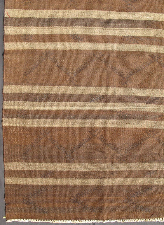 This vintage Tribal Turkish Kilim carpet is set on a natural brown background and features a trio of taupe stripes. The zig-zag pattern in darker brown imbues the piece with a very modern look. This Kilim could function well in a traditional or