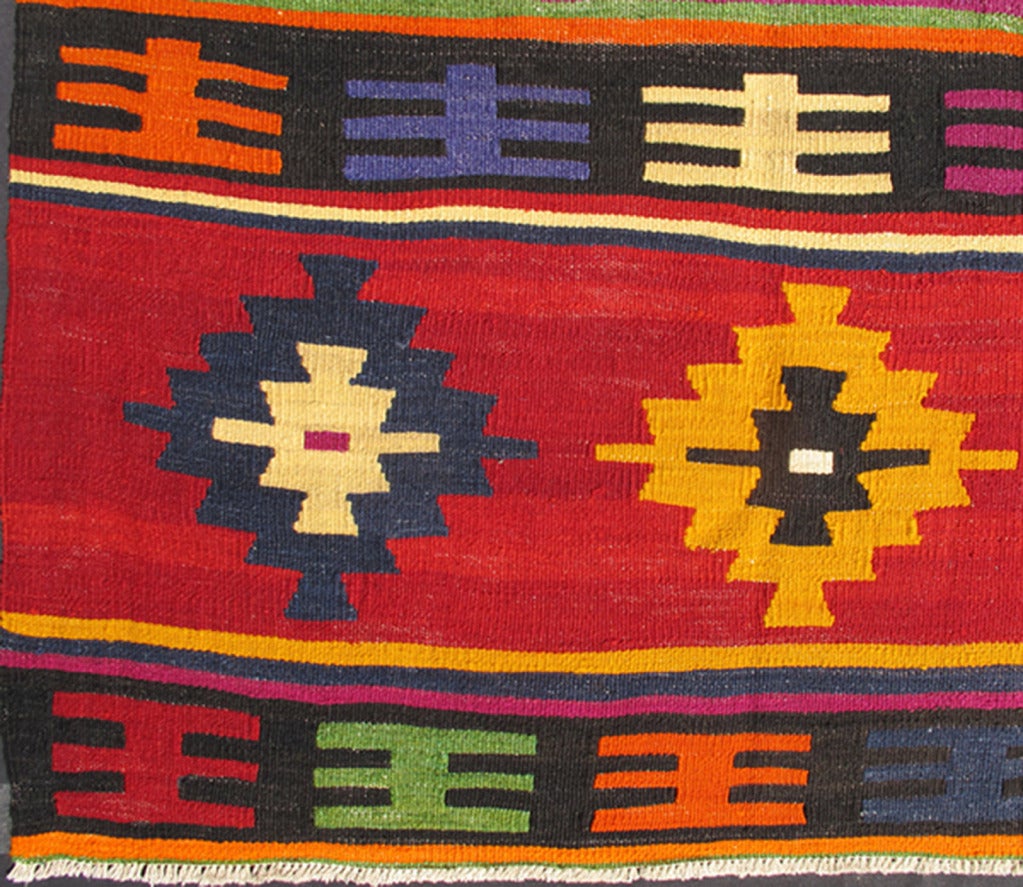 This brilliant and colorful Turkish Kilim rug has a repeating diamond pattern, which is articulated on a background of thick bands of alternating color. This piece is vibrant with wonderfully bright colors, including orange, green, purple, yellow,