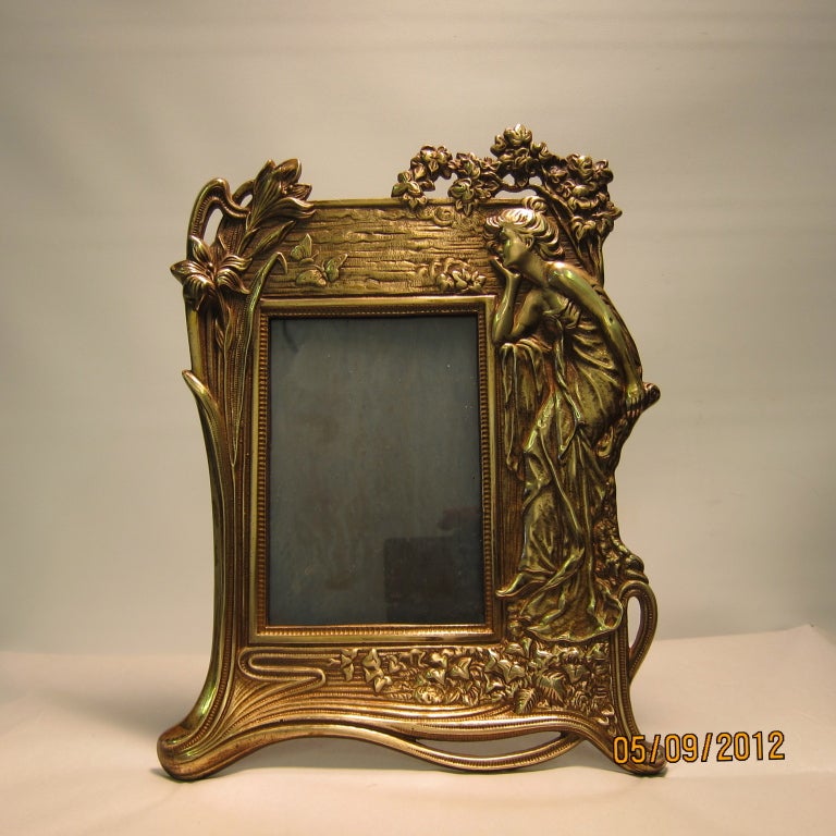 A pair of vintage Art Nouveau bronze photo frames, each signed with intertwined
initials. One frame displays a stylish young woman with flowing hair paying hoagie to an exaggerated flowering iris bloom. She stands prominently against a textured